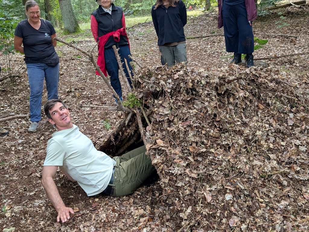 Ein Mann liegt in einer selbstgebauten Laubhütte im Wald bei einem Teamtag.